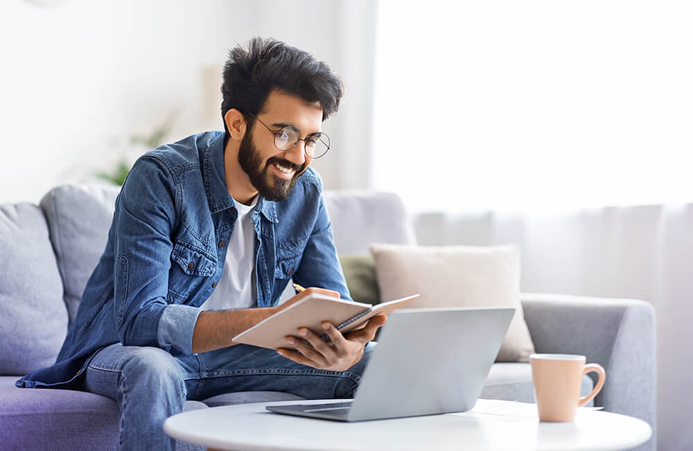 Homem jovem sorridente lendo um livro com um laptop e uma caneca sobre a mesa em uma sala de estar iluminada.