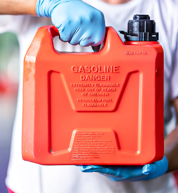 Mãos com luvas segurando um galão de gasolina com etiquetas de advertência.